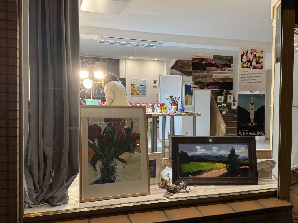 Das Atelier von Roswitha Schumacher-Kuckelkorn leuchtet in die Nacht wie ein offenes Versprechen. Die große Scheibe, einst Schaufenster, ist jetzt ein Rahmen – kein Abstand mehr zwischen Straße und Kunst. Passanten bleiben stehen, gebannt vom Licht, das über die Farben streicht, sie lebendig macht.
Drinnen steht Roswitha, umgeben von Pinseln, Gläsern, Tuben, Leinwänden. Auf dem Tisch eine kleine Ordnung im Chaos der Ideen. Rot, Grün, Blau – jede Farbe will zuerst gehört werden. Im Vordergrund: Tulpen in einem Glas, kräftig und verletzlich zugleich. Daneben eine weite Landschaft, in der Licht über Wiesen läuft, als wüsste es den Weg.
Das Atelier ist Bühne und Werkstatt zugleich. Hier geschieht Malerei im Moment, sichtbar, ungeschützt. Die große Scheibe macht sie öffentlich, ohne sie zu entzaubern. Man spürt, dass Roswitha nicht malt, um etwas zu zeigen – sie malt, um etwas zu finden.
Rheinstrasse 207 | Das Atelier von Roswitha Schumacher-Kuckelkorn | 2025